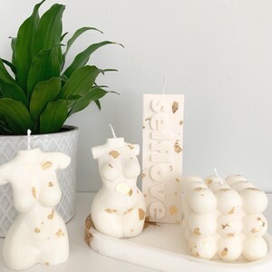 May include: Four white candles with gold flecks. Two candles are shaped like a torso, one is a rectangular pillar with the word "selflove" embossed on it, and the last is a cube-shaped candle with a bubble texture.