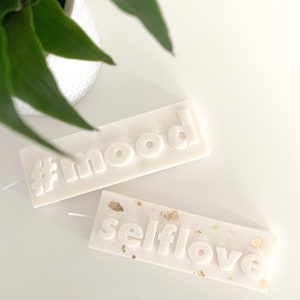 May include: Two white pillar candles with the words "#mood" and "selflove" embossed on them. The "selflove" candle has gold flecks on the surface.