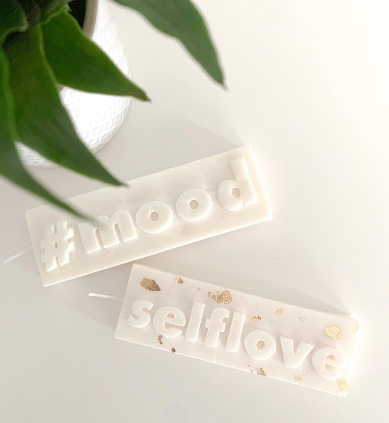 May include: Two white pillar candles with the words "#mood" and "selflove" embossed on them. The "selflove" candle has gold flecks on the surface.