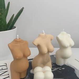 May include: Three nude-toned body shaped candles and one white round candle with a textured surface. The candles are arranged on a book with the word "CHANEL" printed on the cover.
