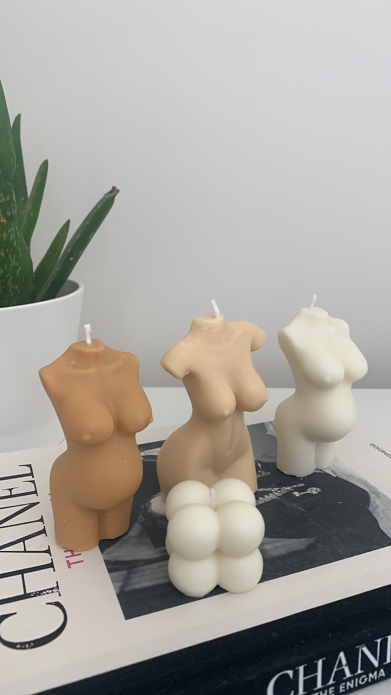 May include: Three nude-toned body shaped candles and one white round candle with a textured surface. The candles are arranged on a book with the word "CHANEL" printed on the cover.