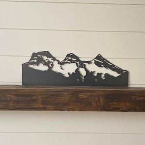 May include: A black metal mountain range silhouette decorates a dark brown wooden mantel above a black fireplace.