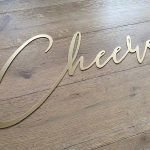 May include: Gold metal wall decor in the shape of the word "Cheers" on a wooden background.