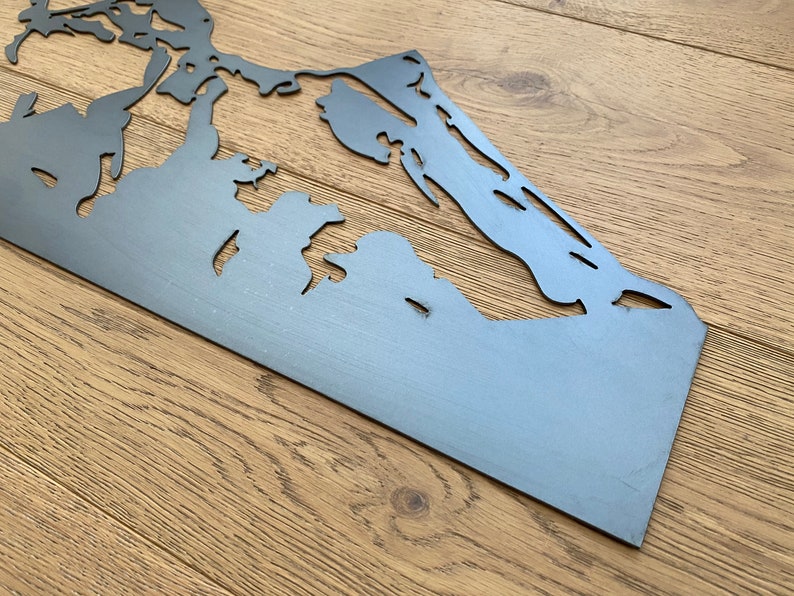 May include: A metal cutout of a mountain range, with a gray finish. The cutout is on a wooden surface.