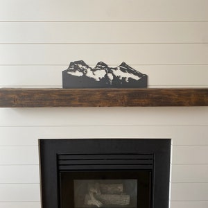 May include: A black metal mountain range silhouette wall decor above a fireplace with a black mantel and a black fireplace insert with a glass door and logs inside.