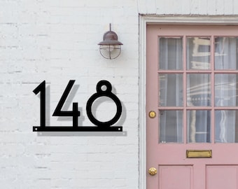 May include: Black metal house number sign displaying the number 148. The numbers are in a modern font and mounted on a white brick wall. A pink door is visible to the right.