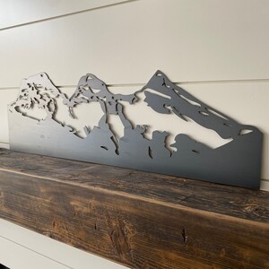 May include: A metal silhouette of a mountain range, painted in a dark gray color, is mounted on a dark brown wooden mantel.