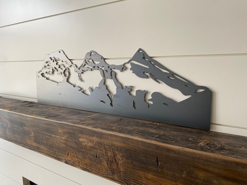 May include: A metal silhouette of a mountain range, painted in a dark gray color, is mounted on a dark brown wooden mantel.