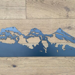May include: A black metal silhouette of a mountain range, with three peaks, against a woodgrain background.