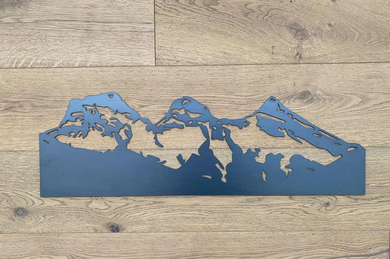 May include: A black metal silhouette of a mountain range, with three peaks, against a woodgrain background.