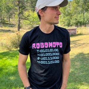 Peut inclure: Un t-shirt noir avec un motif rose et bleu qui dit "ROBOMOTO" et une série de chiffres. La personne portant le t-shirt porte une casquette de baseball blanche avec un logo de taureau rouge.