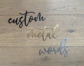 May include: Three metal words, "custom", "metal", and "words", are displayed on a woodgrain background. The words are in a cursive font and are different colors: black, gold, and silver.