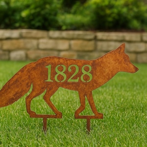 May include: A rusty brown metal fox garden stake with the number 1828 cut out in green. The fox silhouette is standing on two stakes, placed in green grass. The background includes a stone wall and green foliage.