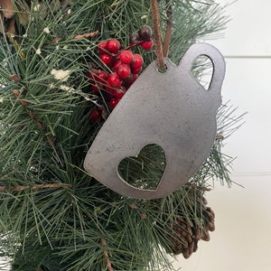 May include: A rustic metal Christmas ornament shaped like a coffee mug with a heart cut out. The ornament is hanging on a green artificial Christmas tree.