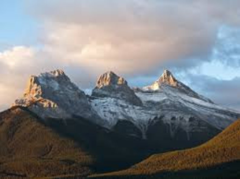 May include: Three snow-capped mountain peaks rise against a cloudy sky, with a forest in the foreground.