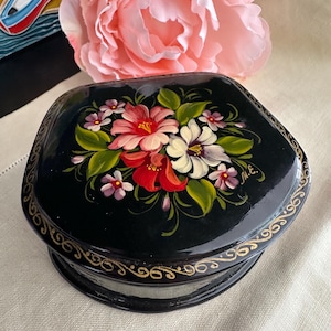 May include: A black lacquered trinket box with a hand-painted floral arrangement featuring pink, red, and white blossoms with green leaves on the lid. The edges of the lid are decorated with a delicate gold filigree pattern, and the initials M.E. are painted in gold near the flowers.