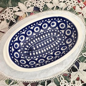 May include: An oval ceramic serving dish with a white rim and a blue and white pattern. The pattern features concentric circles and dotted lines. The dish is placed on a white lace doily.