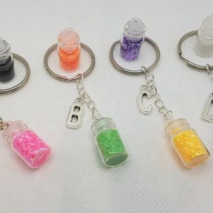 Könnte beinhalten: Schlüsselanhänger mit kleinen Glasflaschen, gefüllt mit bunten Glitzer. Jede Flasche ist an einem silbernen Schlüsselring und einem Anhänger mit einem Buchstaben befestigt. Die Glitzerfarben umfassen Rosa, Grün, Gelb und Blau.