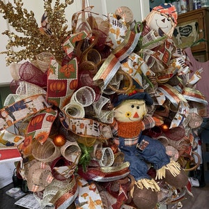 May include: A fall-themed wreath with burlap, plaid, and brown and white patterned ribbon. Two scarecrows are attached to the wreath, one with a red and white hat and the other with a brown hat.