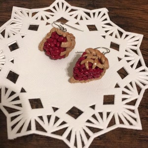 Può includere: Un paio di orecchini pendenti con una crosta di torta in miniatura piena di frutta rossa. Gli orecchini sono realizzati in pasta polimerica e hanno ganci d'argento.