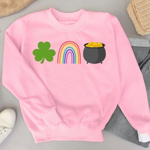 Pode incluir: Camisola cor-de-rosa com um trevo verde, um arco-íris e um pote de ouro.