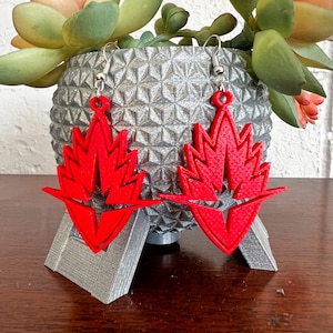 May include: A pair of red dangle earrings with a geometric design. The earrings are made of a lightweight material and have a silver hook.