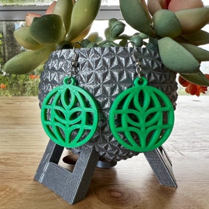 May include: A pair of green earrings with a geometric design. The earrings are hanging from a silver stand.