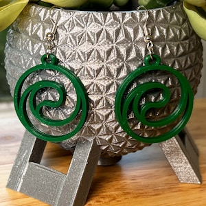 May include: A pair of green dangle earrings with a swirling design. The earrings are made of a lightweight material and have silver hooks.