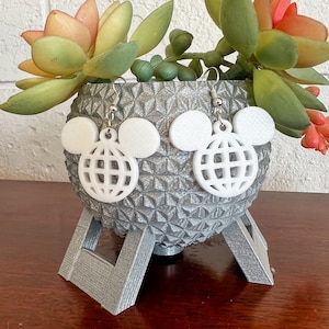 May include: White Mickey Mouse-shaped earrings with a white globe design in the centre. The earrings are displayed on a silver, textured, sphere-shaped stand.