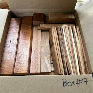 May include: A cardboard box labeled Box #7 containing a collection of rectangular wooden pieces in various shades of brown. The assortment includes several long, flat planks with visible wood grain patterns and smaller, narrow strips of wood.
