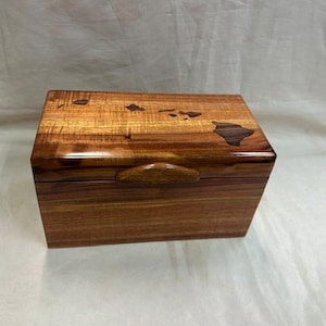 May include: A rectangular wooden box with a glossy finish. The box is made of light and dark brown wood, with a handle on the front. The top of the box features an inlay of the Hawaiian Islands. The box is sitting on a white surface.