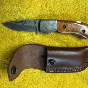 May include: A folding knife with a patterned blade and a wooden handle, next to a brown leather sheath. The knife blade is dark with a unique pattern. The handle is a light brown color with a wood grain pattern.