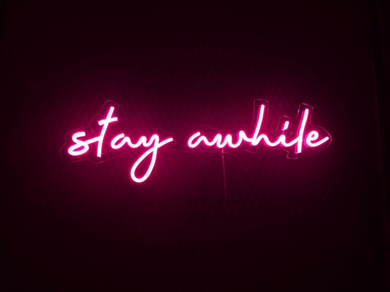 K&ouml;nnte beinhalten: Rosa leuchtendes Neon-Schild mit dem Text "stay awhile" auf schwarzem Hintergrund.