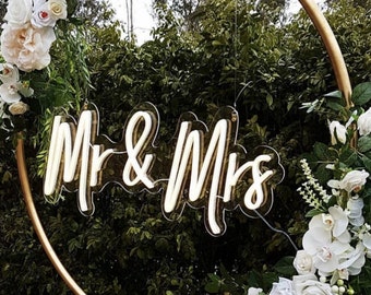 May include: White neon sign with the words "Mr & Mrs" hanging from a gold hoop decorated with white flowers.