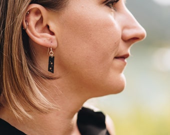 May include: A woman wearing a black shirt with a pair of black dangle earrings. The earrings have a rectangular shape with small white dots.