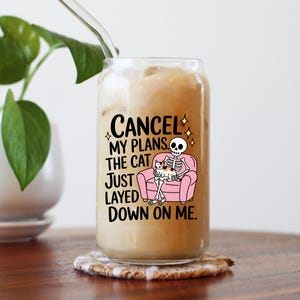May include: A clear glass tumbler, shaped like a can, filled with a creamy drink and a glass straw. The tumbler has a graphic of a skeleton on a pink sofa with a cat, and the text "CANCEL MY PLANS THE CAT JUST LAYED DOWN ON ME."