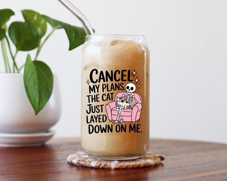 May include: A clear glass tumbler, shaped like a can, filled with a creamy drink and a glass straw. The tumbler has a graphic of a skeleton on a pink sofa with a cat, and the text "CANCEL MY PLANS THE CAT JUST LAYED DOWN ON ME."
