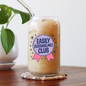 K&ouml;nnte beinhalten: Ein klares, dosenf&ouml;rmiges Glas, gef&uuml;llt mit einem hellbraunen Getr&auml;nk und Eis. Das Glas hat ein lila und rosa Abzeichen mit der Aufschrift "EASILY OVERWHELMED CLUB". Ein klarer Glasstrohhalm steckt in dem Getr&auml;nk. Das Glas steht auf einem gemusterten Untersetzer.