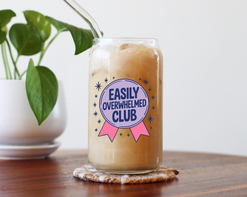 K&ouml;nnte beinhalten: Ein klares, dosenf&ouml;rmiges Glas, gef&uuml;llt mit einem hellbraunen Getr&auml;nk und Eis. Das Glas hat ein lila und rosa Abzeichen mit der Aufschrift "EASILY OVERWHELMED CLUB". Ein klarer Glasstrohhalm steckt in dem Getr&auml;nk. Das Glas steht auf einem gemusterten Untersetzer.