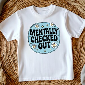 以下が含まれることがあります： レトロなデザインの白いTシャツ。デザインは、黒で「MENTALLY CHECKED OUT」というテキストを、白いデイジーと黄色の星のアクセントが付いた水色の円の中に配置しています。シャツは柔らかい素材で作られています。