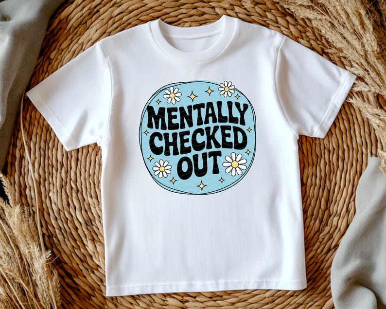 以下が含まれることがあります： レトロなデザインの白いTシャツ。デザインは、黒で「MENTALLY CHECKED OUT」というテキストを、白いデイジーと黄色の星のアクセントが付いた水色の円の中に配置しています。シャツは柔らかい素材で作られています。