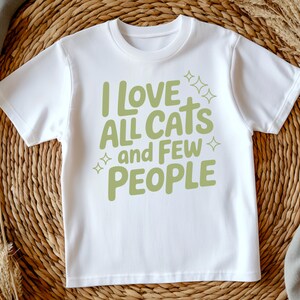 May include: White t-shirt featuring the phrase "I LOVE ALL CATS and FEW PEOPLE" in light green, with star motifs. The text is in a casual font. The shirt is presented on a woven surface.
