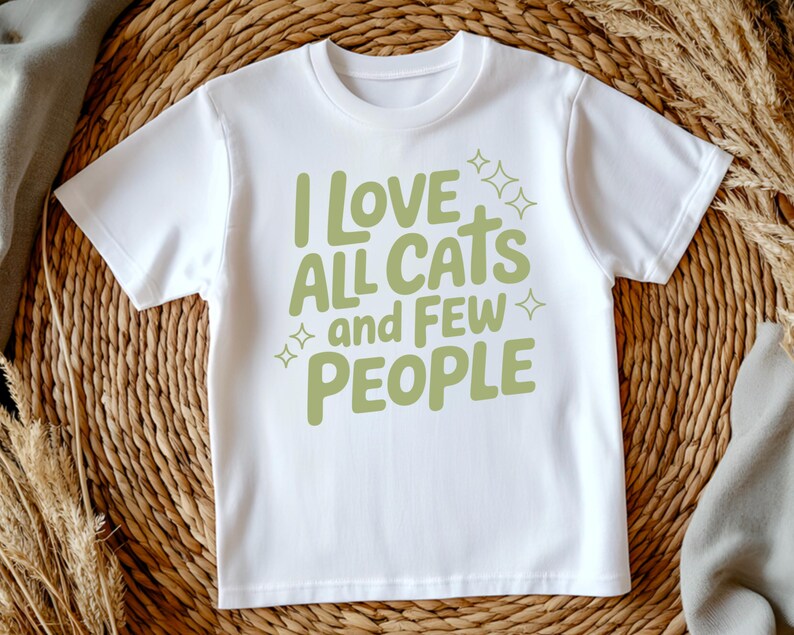 May include: White t-shirt featuring the phrase "I LOVE ALL CATS and FEW PEOPLE" in light green, with star motifs. The text is in a casual font. The shirt is presented on a woven surface.