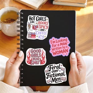 May include: A black notebook with four colorful stickers. The stickers have text that reads: "Hot Girls Like Books Spicy and Coffee Icy", "Good Girls Read Smut", "I'm Looking For a Man Written by a Woman", and "Feral over Fictional Men".