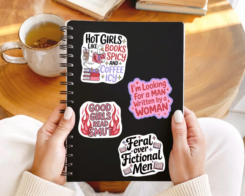 May include: A black notebook with four colorful stickers. The stickers have text that reads: "Hot Girls Like Books Spicy and Coffee Icy", "Good Girls Read Smut", "I'm Looking For a Man Written by a Woman", and "Feral over Fictional Men".