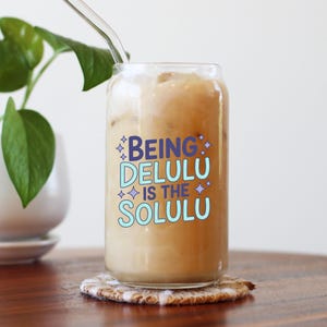 May include: A clear glass tumbler filled with a light brown beverage and ice, with a glass straw. The text "BEING DELULU IS THE SOLULU" is printed on the glass in blue and light blue. The glass sits on a patterned coaster.