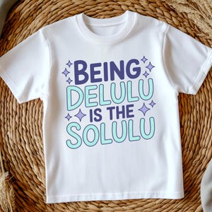 May include: White t-shirt with the text "BEING DELULU IS THE SOLULU" in purple and light blue. The text is surrounded by small star shapes. The shirt is made of a soft material.