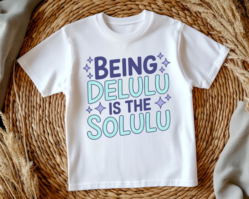 May include: White t-shirt with the text "BEING DELULU IS THE SOLULU" in purple and light blue. The text is surrounded by small star shapes. The shirt is made of a soft material.