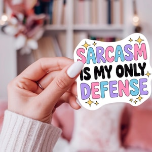 May include: A white sticker with the text "SARCASM IS MY ONLY DEFENSE" in colorful bubble letters. The sticker has a unique shape and is decorated with yellow star accents. The sticker is held by a person.