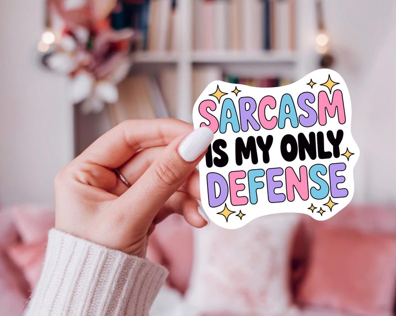 May include: A white sticker with the text "SARCASM IS MY ONLY DEFENSE" in colorful bubble letters. The sticker has a unique shape and is decorated with yellow star accents. The sticker is held by a person.